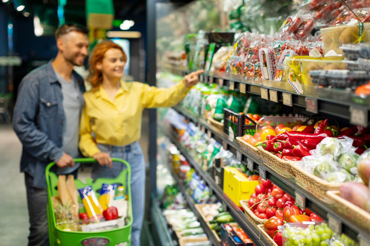 Grocery shopping. Young couple choosing groceries in supermarket, selective focus on fresh vegetables on shelf