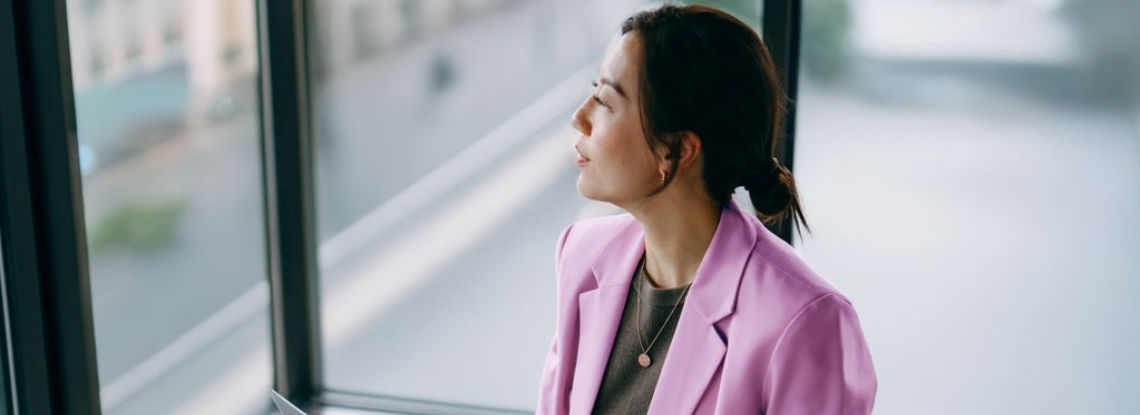 A woman is sat looking out of a hotel window with a laptop on her knee.