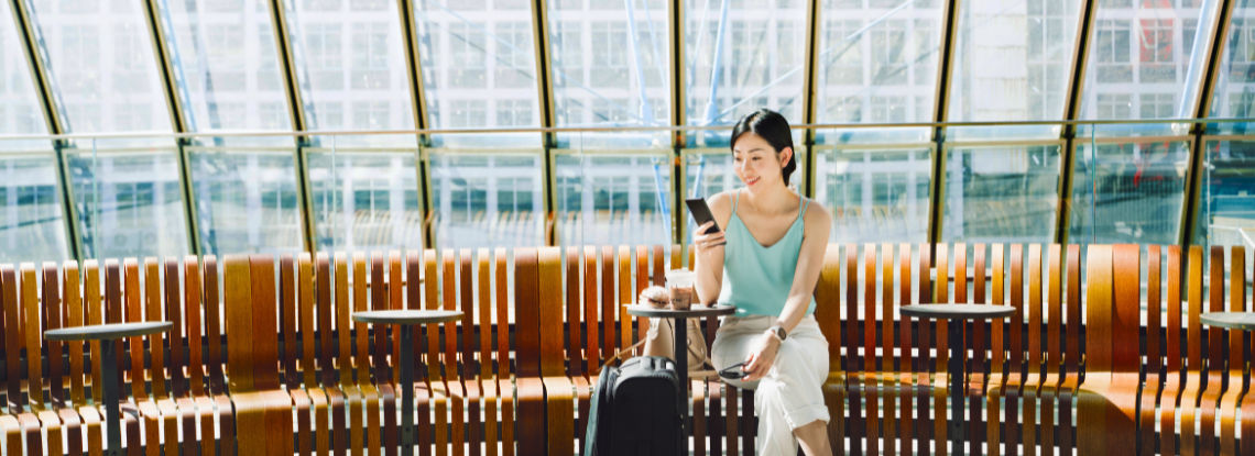 A woman in a blue top smiles at her phone while she sits on a bench in a station in front of a large window with a suitcase.