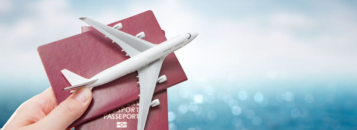 A hand holding up two passports behind a plane replica on a background with a blue sky and the ocean.