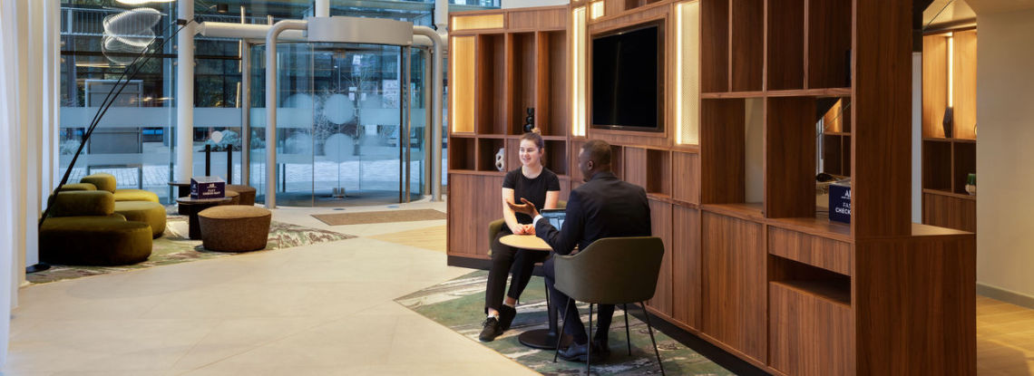 A man and a woman sit with laptops in a modern hotel lobby, engaged in conversation near wooden shelves and a large window.