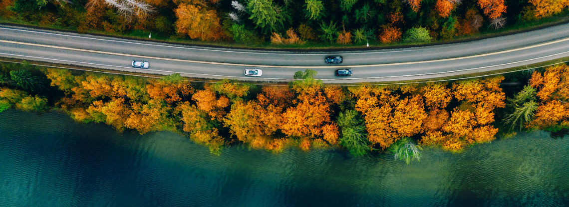 Aerial view of a road bordered by autumn trees and a blue lake, with cars driving along the route.