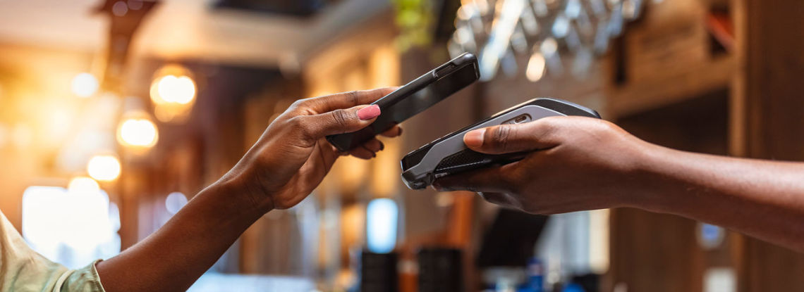 A hand holding a phone to make a contactless payment on a card reader held by another person.