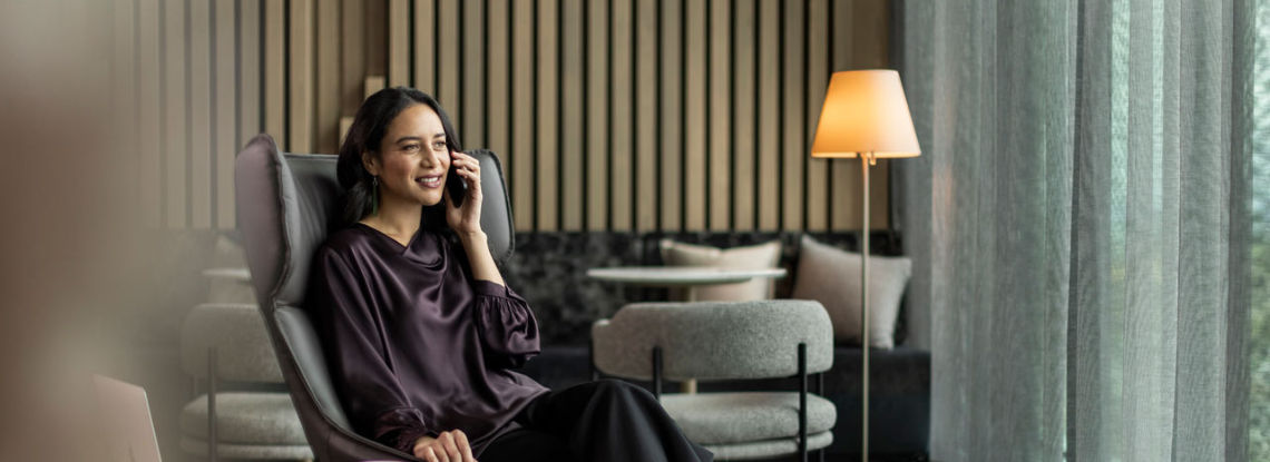 A woman in smart attire sits in a modern lounge chair in a hotel lobby, smiling while talking on the phone.