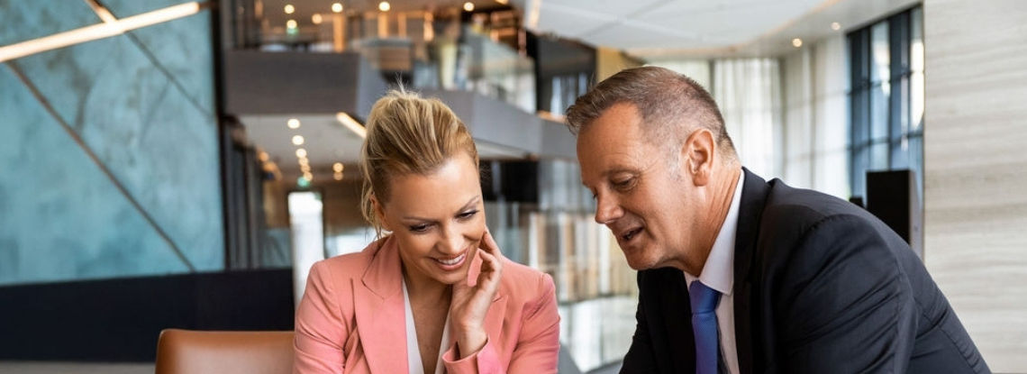 A woman and man sit on chairs, the man is holding an iPad and they are both looking at the screen.