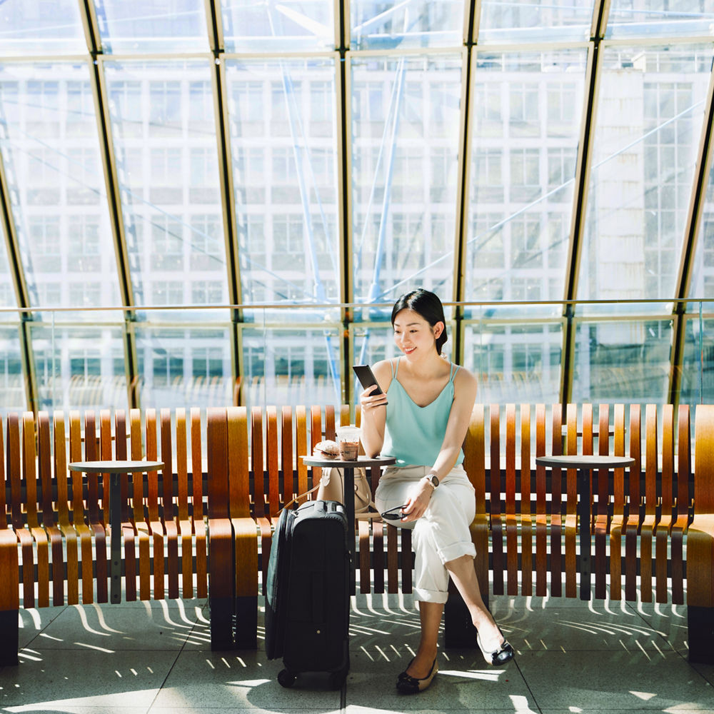 A woman in a blue top smiles at her phone while she sits on a bench in a station in front of a large window with a suitcase.