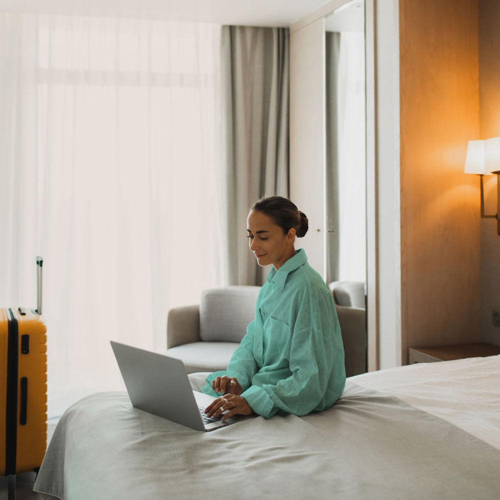 A woman is sitting on a bed in a hotel room looking on her laptop.