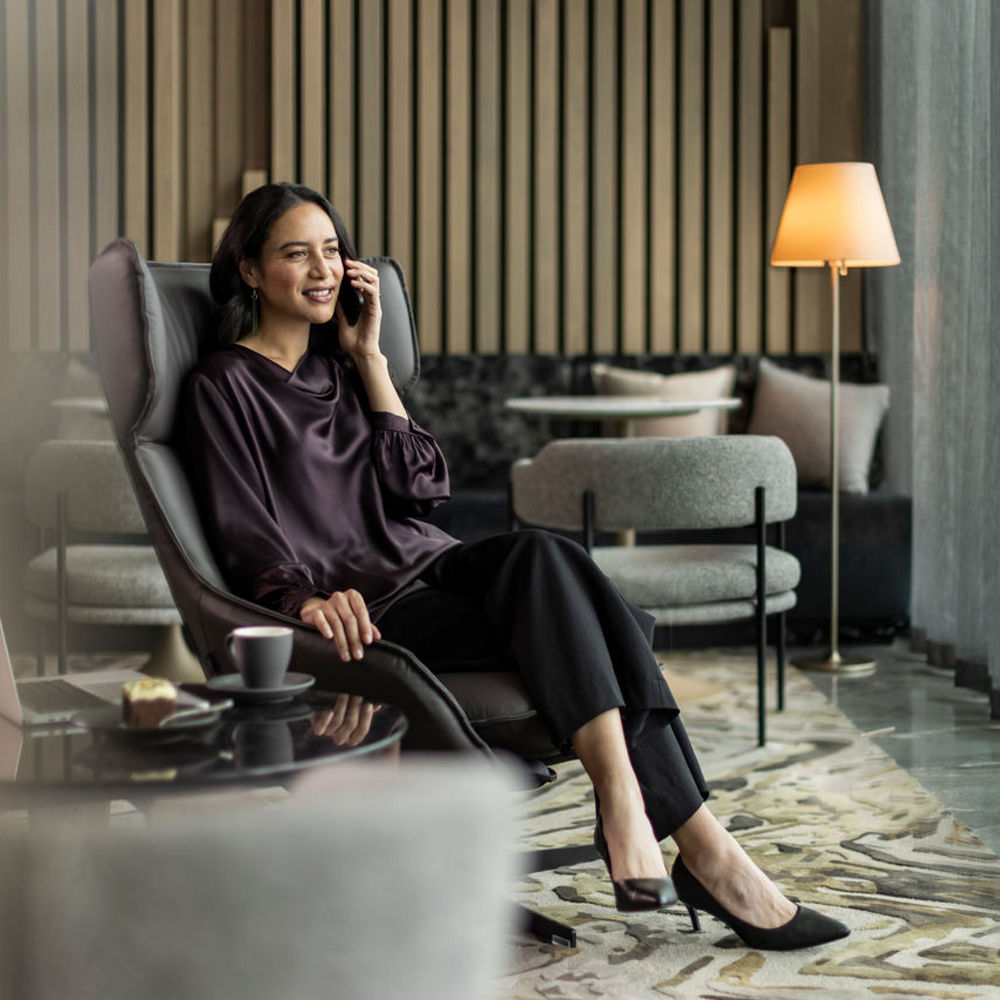 A woman in smart attire sits in a modern lounge chair in a hotel lobby, smiling while talking on the phone.