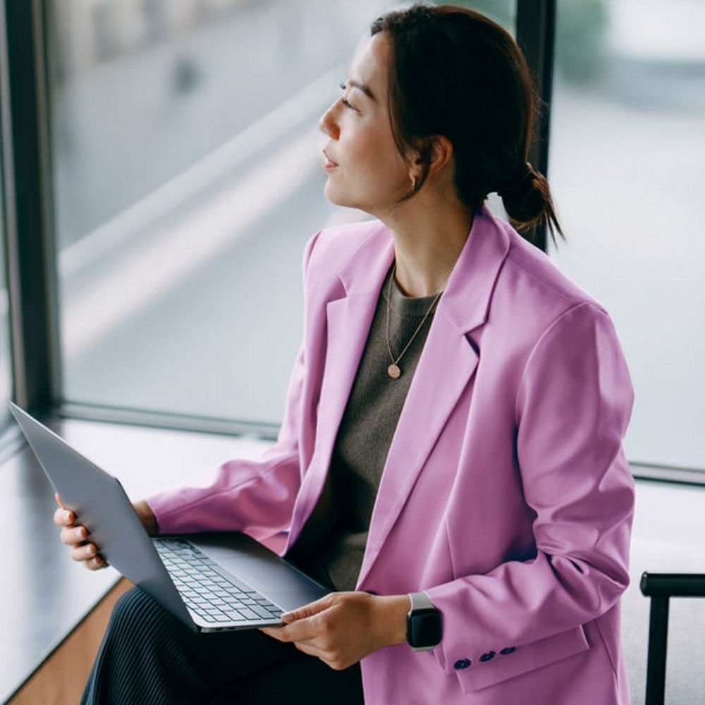 A woman is sat looking out of a hotel window with a laptop on her knee.