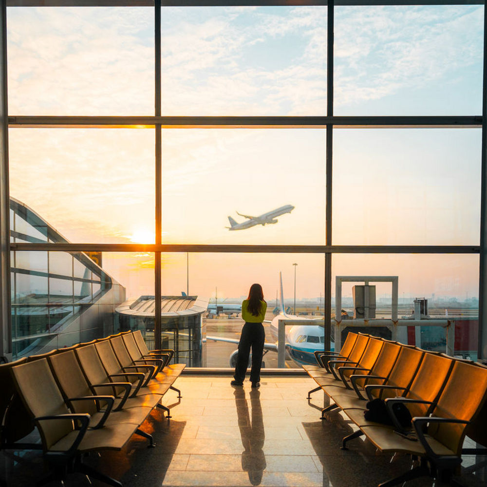 A woman standing in an empty airport lounge looking out of the window at a plane taking off.