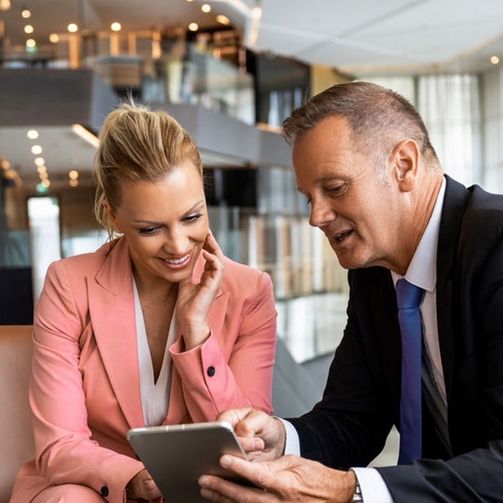 A woman and man sit on chairs, the man is holding an iPad and they are both looking at the screen.