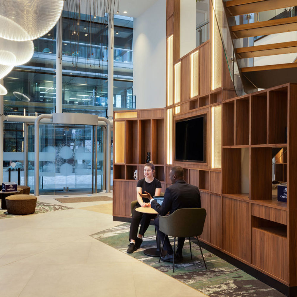 A man and a woman sit with laptops in a modern hotel lobby, engaged in conversation near wooden shelves and a large window..