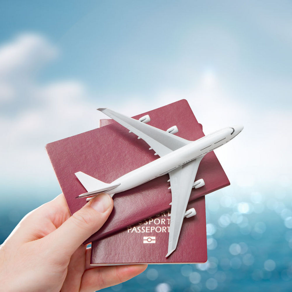 A hand holding up two passports behind a plane replica on a background with a blue sky and the ocean.