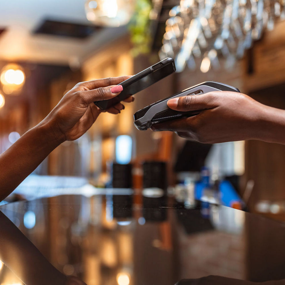 A hand holding a phone to make a contactless payment on a card reader held by another person.