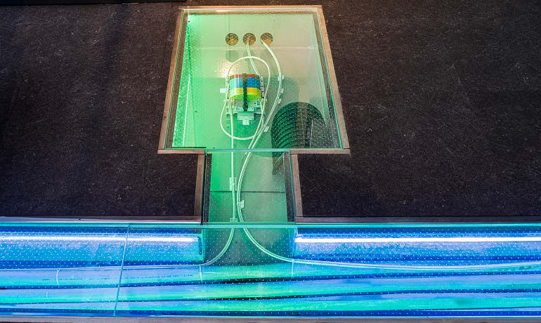 Sezione espositiva con pavimento trasparente che mostra cavi elettrici e tubature illuminate da luci LED colorate