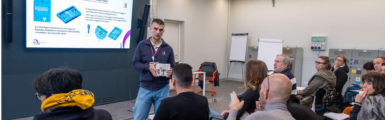 Una classe di persone assiste ad una lezione. Si intravede una slide proiettata e c'è un docente che parla tenendo in mano uno strumento di lavoro, oggetto della lezione