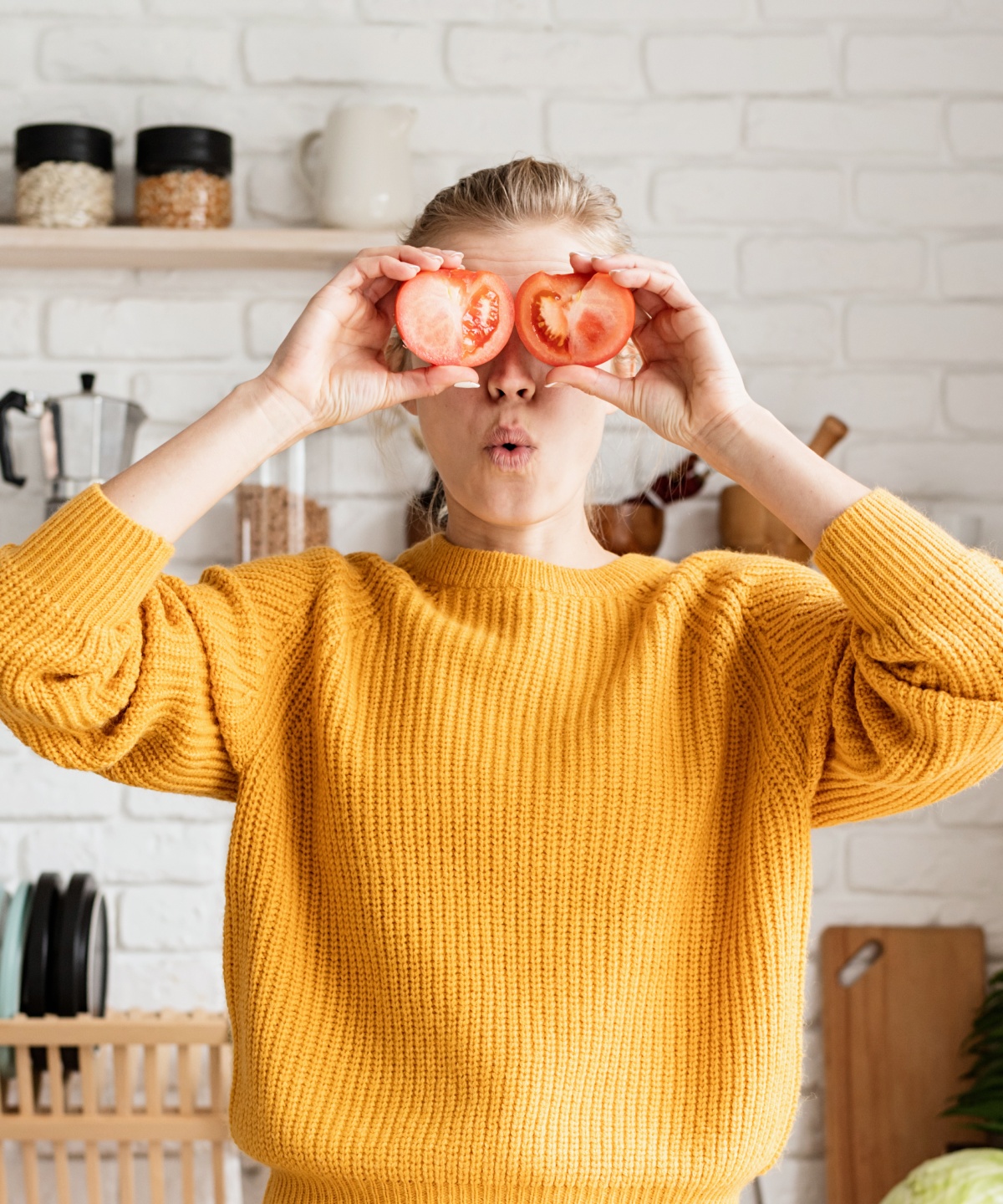 Junge Frau mit gelben Woll-Pullover hält sich zwei Tomatenhälften vor die Augen.