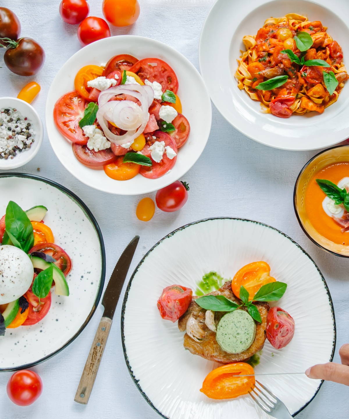 Draufsicht eines Tisches mit weißer Tischdecke und vielen Tellern und Schalen, die mit aufgeschnittenen Tomaten und verschiedenen Tomatenspeisen gefüllt sind.