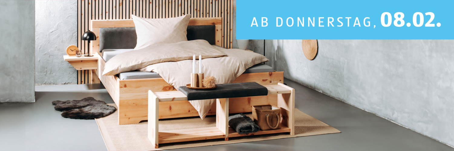 Angebote ab Donnerstag, 08.02. - Abgebildet ist ein Bett aus Holz mit hellbrauner Bettwäsche in einem Raum mit grauen Wänden.