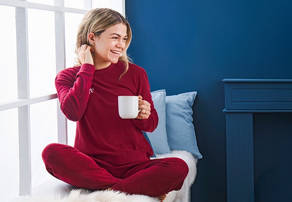 Ženska v rdeči pižami sedi ob oknu, v roki ima skodelico