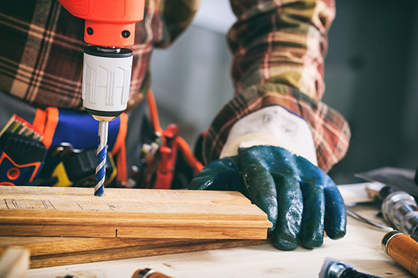 Eine Mann hält mit der linken Hand ein Brett. Mit der anderen benutzt er einen Bohrer. Er trägt Arbeitshandschuhe.