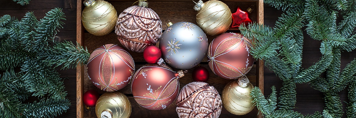 Sehr fein verzierte Christbaumkugeln in Rosa, Blau und Gold liegen in einer Holzkiste, daneben sind Tannenzweige.