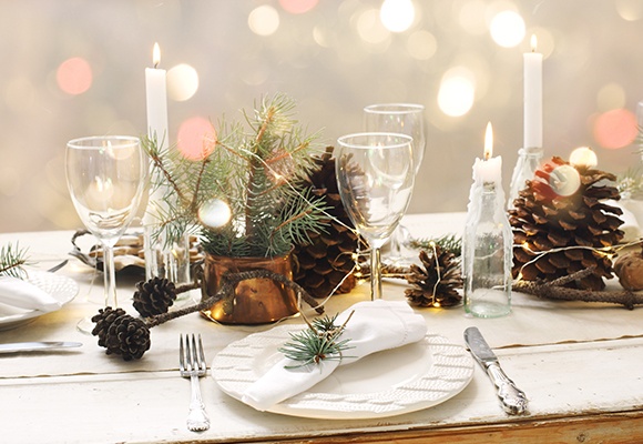 Ein festlich Gedeckter Tisch in Weiß mit leeren Weingläsern, Tannenzweigen, Tannenzapfen und Kerzen in kleinen Flaschen.