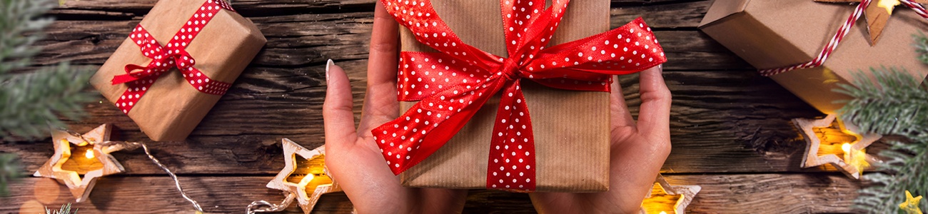 Frauenhände halten ein in braunem Papier eingewickeltes Geschenk mit rot-weißer Schleife. Im Hintergrund sind weitere Geschenke und sternenförmige Kerzen.