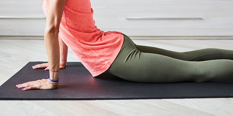 Frau liegt auf einer schwarzen Yogamatte und streckt den Oberkörper durch die Arme nach oben.