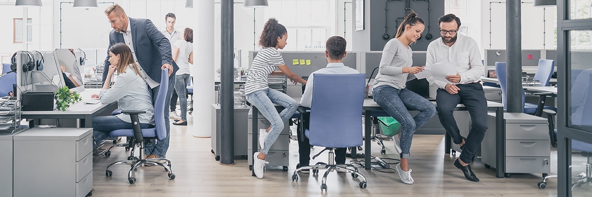 In einem sehr sterilen Büro mit blauen Bürostühlen sind mehrere Mitarbeiter, die gemeinsam diskutieren und arbeiten.