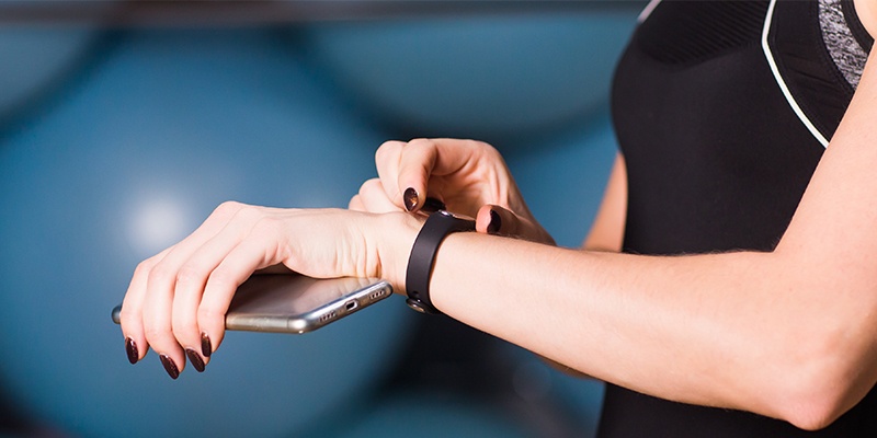 Eine Frau im schwarzen Top hält in der einen Hand ein silbernes Smartphone und stellt mit der anderen Hand ein Fitnessarmband ein. Der Hintergrund ist blau.