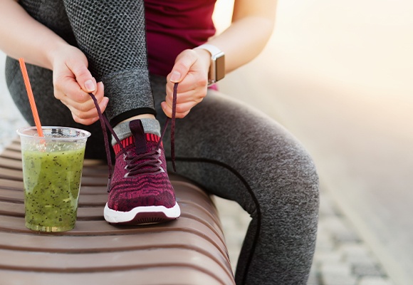 Frau bindet ihren lila-pinken Sportschuh im Sitzen zu, während ein grüner Smoothie vor ihr auf der Bank steht.