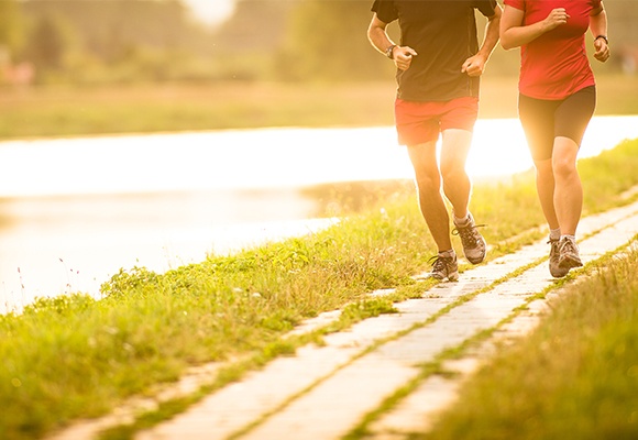 Ein Mann und eine Frau joggen auf einem schmalen Pfad neben einem See im Grünen. Die Sonne geht unter und spiegelt sich im Wasser.