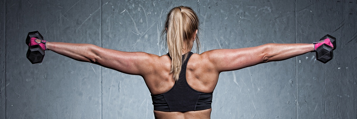 Muskulöse Frau trägt ein schwarzes Sporttop, streckt die Arme aus und hält zwei Hanteln mit rosa Handschuhen. Ansicht von hinten.