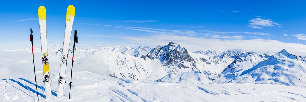 Zwei Skier und Skistöcke stecken im Schnee, während eine Gondel im Hintergrund hochfährt. Der Blick geht über ein Gebirge.