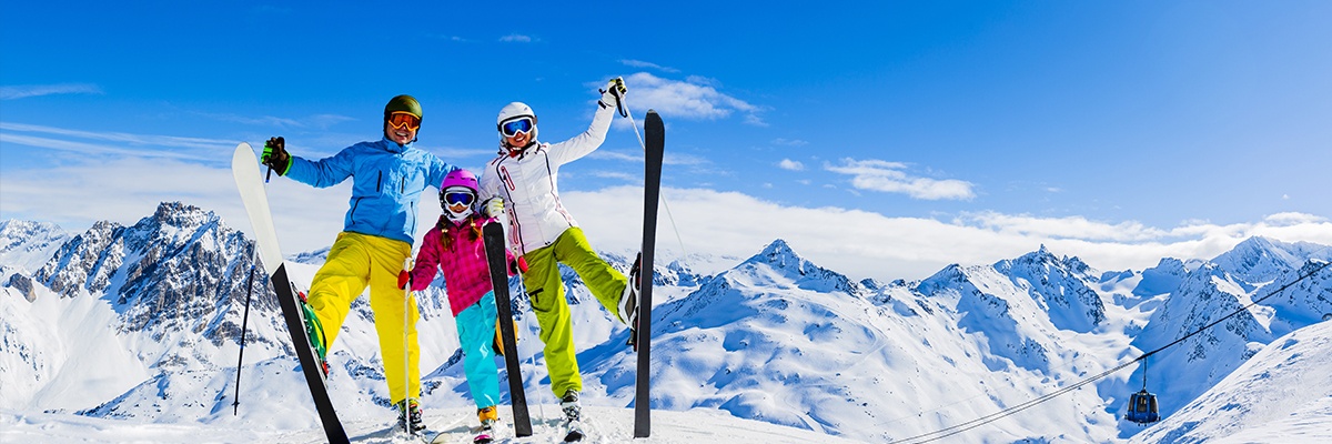 Eine dreiköpfige Familie in bunter Skiausrüstung hebt gemeinsam jeweils ein Bein mit Skiern dran und lacht in die Kamera.