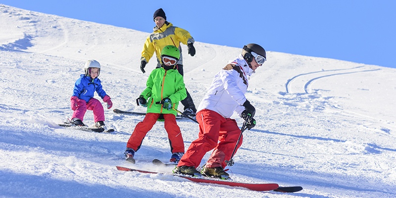 Eine vierköpfige Familie fährt in bunter Skiausrüstung langsam einen relativ flachen Hang herunter.