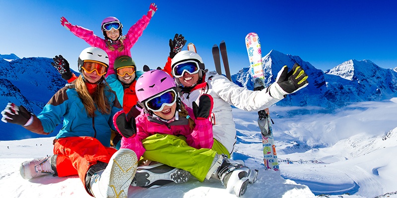 Eine Familie in knallig buntem Outfit steht auf einem verschneiten Berg. Im Hintergrund sieht man Skier und ein weites Gebirge.