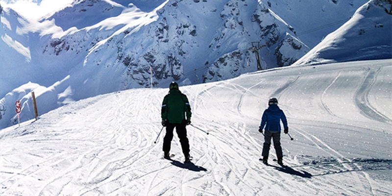 Zwei Skifahrer gleiten einen schwachen Hang bergab, im Hintergrund ist schneebedecktes Gebirge.