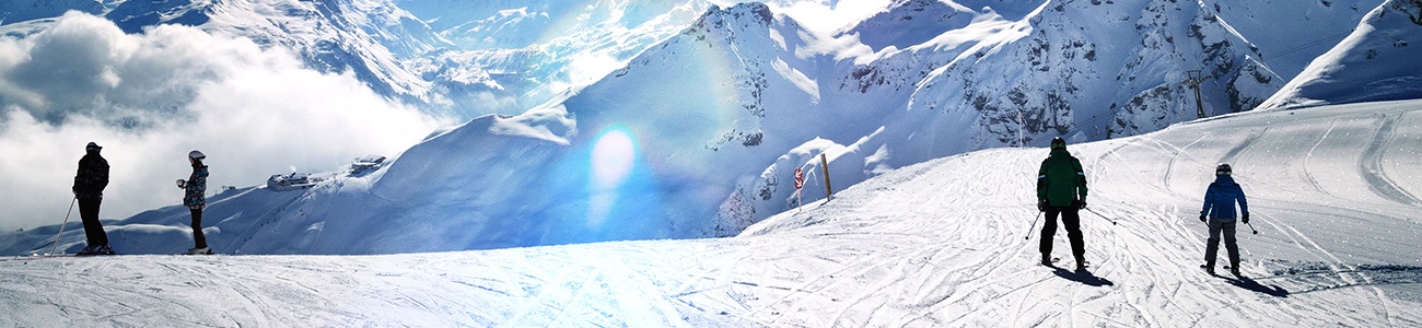 Eine sehr blau leuchtende Berglandschaft wird in Wolken verschluckt, während vier vereinzelte Skifahrer zu sehen sind.