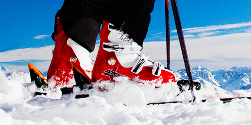 Im Hintergrund ist ein Gebirge, das in blau leuchtet, während vorn zwei rote Skischuhe über Schnee fahren.