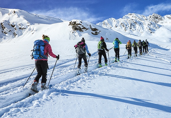 Eine Reihe mehrere Skifahrer folgt einer vorgegeben Spur im Schnee inmitten eines Gebirges.