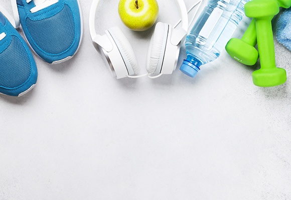 Verschiedene Sportmaterialien wie Joggingschuhe, grüne Hanteln, eine Wasserflasche, Köpfhörer und ein Handtuch liegen auf einem weißen Untergrund.