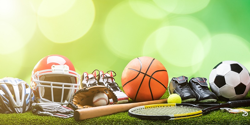 Verschiedene Sportausrüstungen wie Baseballschläger, Federballschläger, Boxhandschuhe, Fußballschuhe, Fahrradhelm und Co liegen auf Kunstrasen.