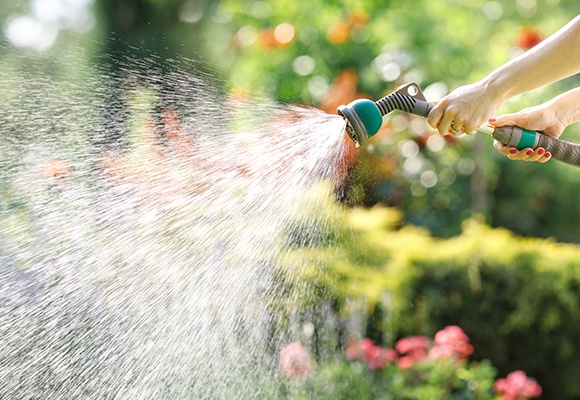 Zwei Frauenhände halten einen türkisen Wasserschlauch mit Aufsatz. Wasser spritzt nach links weg.