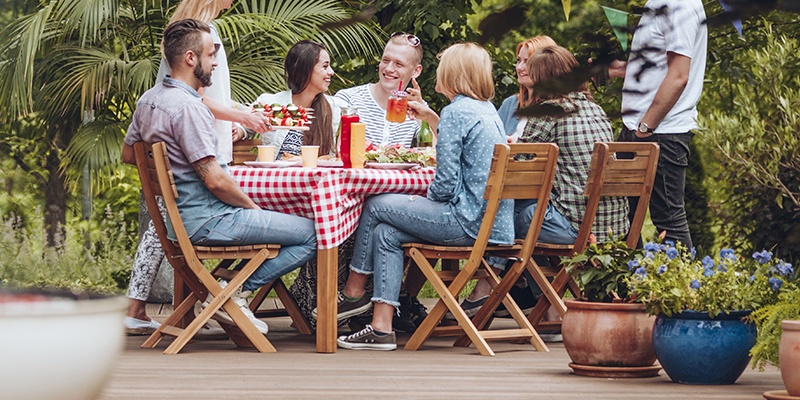 Eine Gruppe Freunde sitzt gemeinsam an einem Gartentisch auf Holzstühlen. Die Tischdecke ist rot-weiß kariert.