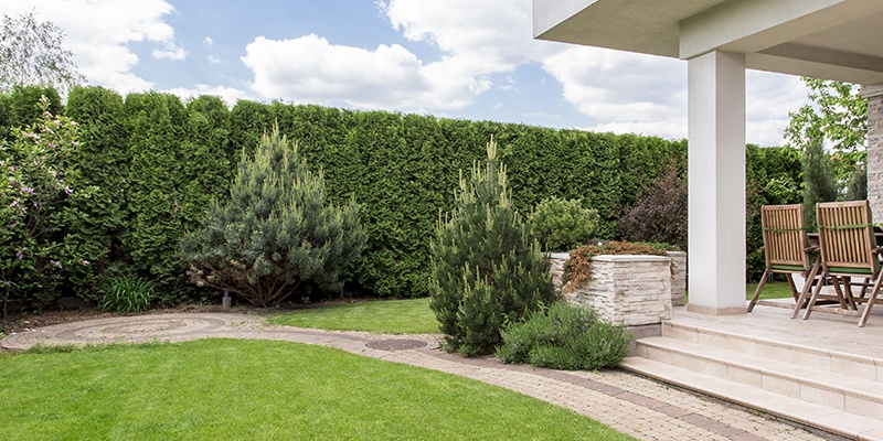 Eine große Hecke umringt eine Terrasse mit Standsäule, runde Wege zieren den sauber geschnittenen Rasen und verschiedene Büsche.