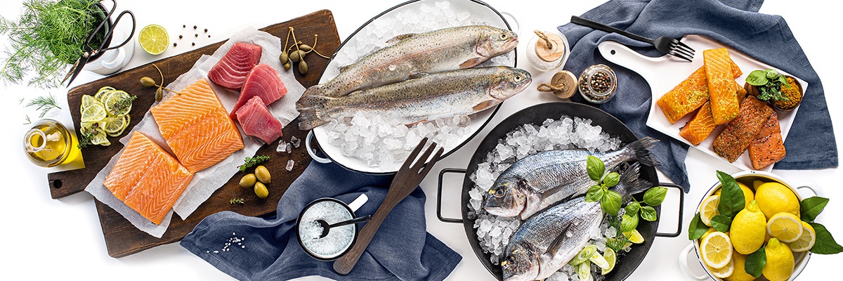 Aufgeschnittener Frisch wie Lachs und Thunfisch liegen auf einem Holzschneidebrett. Ganzer Fisch liegt gekühlt auf Eiswürfeln. Zitronen und Gewürze runden das Bild ab.