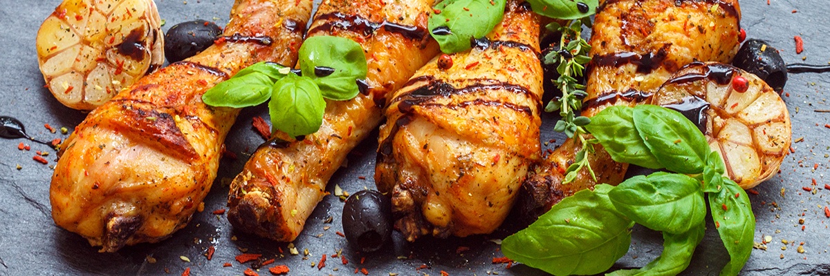 Vier Hühnerkeulen mit goldbrauner Haut liegen auf einer Steinplatte belegt mit Oliven und Kräutern.