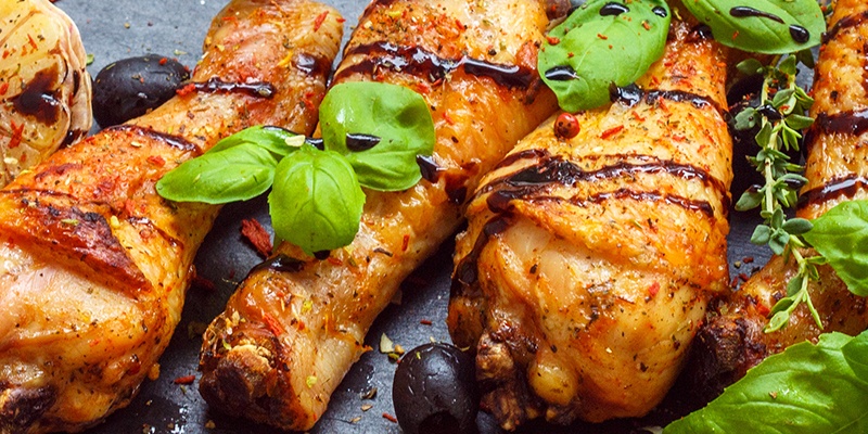 Vier Hühnerkeulen mit goldbrauner Haut liegen auf einer Steinplatte belegt mit Oliven und Kräutern.